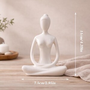 May include: A white ceramic yoga figurine in a seated lotus position. The figurine is 4.33 inches tall and 2.95 inches wide. The background includes a wooden tray with stones, a candle, and a vase with greenery.