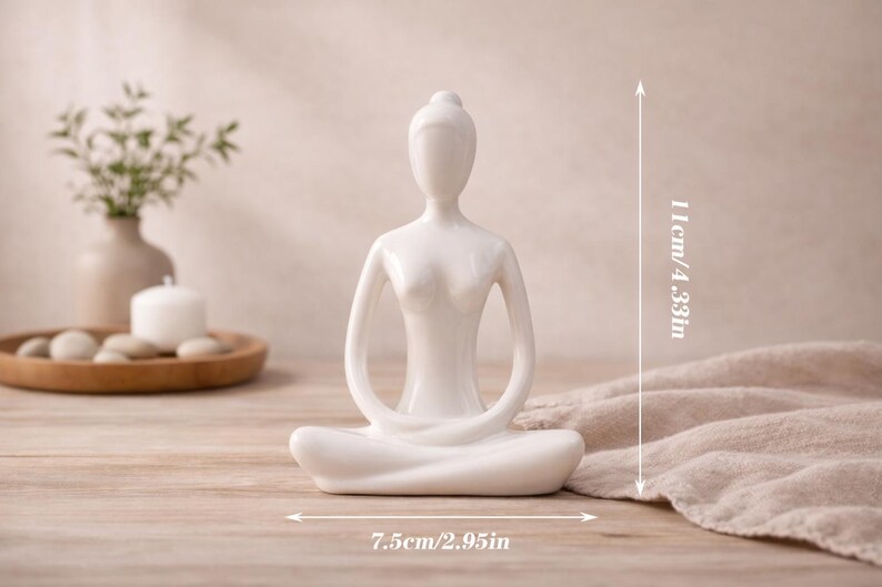 May include: A white ceramic yoga figurine in a seated lotus position. The figurine is 4.33 inches tall and 2.95 inches wide. The background includes a wooden tray with stones, a candle, and a vase with greenery.