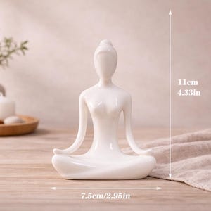 May include: A white ceramic yoga figurine in a seated lotus position. The figurine is 4.33 inches tall and 2.95 inches wide. The figurine is on a wooden surface, with a neutral background. The figurine is a decorative item.