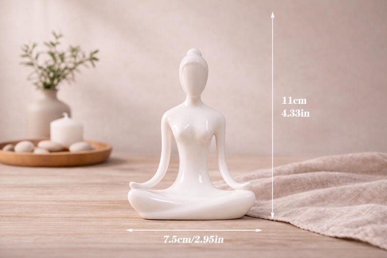 May include: A white ceramic yoga figurine in a seated lotus position. The figurine is 4.33 inches tall and 2.95 inches wide. The figurine is on a wooden surface, with a neutral background. The figurine is a decorative item.