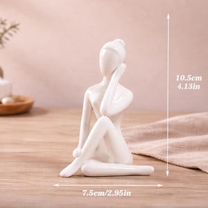 May include: A white ceramic figurine of a seated person, with one arm raised to the head. The figurine is 4.13 inches tall and 2.95 inches wide. The sculpture is on a wooden surface.