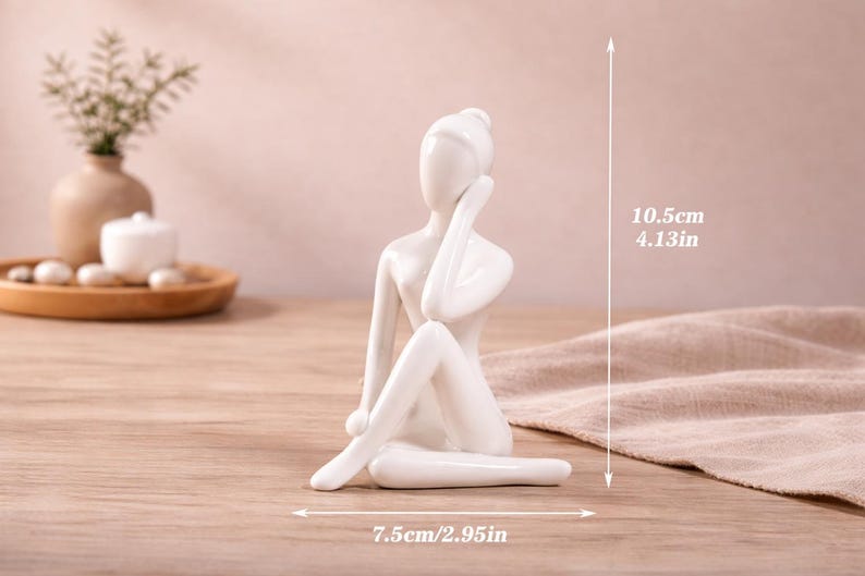 May include: A white ceramic figurine of a seated person, with one arm raised to the head. The figurine is 4.13 inches tall and 2.95 inches wide. The sculpture is on a wooden surface.