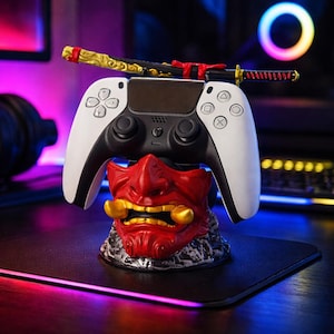 May include: A black and white gaming controller rests on a red and gold Oni mask stand, with a miniature katana on top. The controller has white sides and black center. The stand is detailed with a textured base.