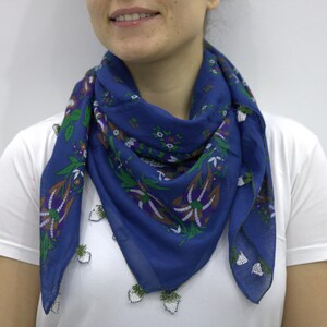 May include: A blue scarf with a floral pattern in green, purple, and white is draped around the neck. The scarf is made of a lightweight fabric and is worn over a white t-shirt. The floral design includes stylized leaves and flowers.