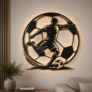 Può includere: Un'opera d'arte da parete circolare in metallo nero che raffigura la silhouette di un calciatore che calcia un pallone, inserita in un grande motivo a pallone da calcio. Il design è retroilluminato con una luce calda che mette in risalto.