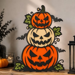 May include: A layered wooden decoration featuring three stacked jack-o'-lanterns with orange and cream-coloured pumpkins, black carved facial features, green leaves, and black bat silhouettes, creating a vertical Halloween-themed display piece.