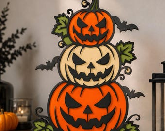 May include: A layered wooden decoration featuring three stacked jack-o'-lanterns with orange and cream-colored pumpkins, black carved facial features, green leaves, and black bat silhouettes, creating a vertical Halloween-themed display piece.