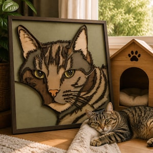 May include: A square framed wall art piece featuring a multi-layered wooden cutout of a tabby cat face with green eyes and brown markings, set against a sage green background, positioned next to a small wooden pet house with a paw print cutout.