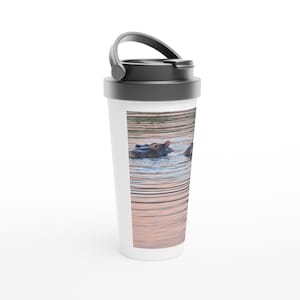 Könnte beinhalten: Ein weißer Reisebecher mit schwarzem Griff und Deckel. Der Becher zeigt ein Foto von zwei Flusspferden im Wasser. Das Wasser hat einen rosafarbenen Farbton, und die Flusspferde sind teilweise untergetaucht. Der Becher ist für heiße oder kalte Getränke konzipiert.