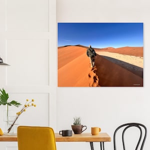 Könnte beinhalten: Ein Wandbild, das eine Person in einer dunklen Jacke und heller Hose zeigt, die auf dem Kamm einer leuchtend orangefarbenen Sanddüne unter einem klaren blauen Himmel geht, mit sichtbaren Fußspuren im Sand und einer weiten Wüstenlandschaft.