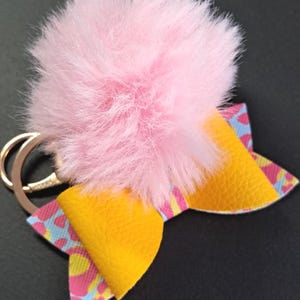 Puede incluir: Un llavero con pompón rosa y anilla dorada. El llavero presenta un lazo amarillo con un estampado colorido de formas rosas, azules y amarillas.