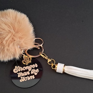 Peut inclure: Un porte-clés avec un pompon duveteux brun clair, des éléments métalliques dorés et un gland blanc. Un charm circulaire noir avec l'inscription "Stronger than the storm" est également attaché. Un petit charm en forme de couronne dorée est également présent.