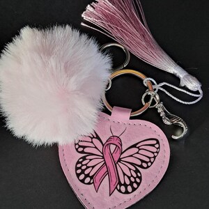 Pode incluir: Um chaveiro rosa em forma de coração com um desenho de borboleta e uma fita rosa, simbolizando a conscientização sobre o câncer de mama. O chaveiro inclui um pompom rosa fofo e uma borla rosa. O hardware de metal é prateado.