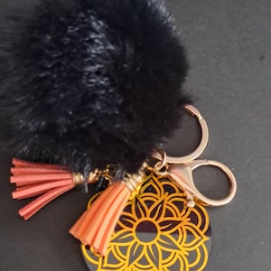 May include: A black keychain with a fluffy pom-pom, peach-coloured tassels, and a gold-coloured circular charm. The charm features a yellow floral design. The keychain has a gold-coloured clasp.
