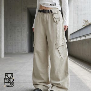 Può includere: Pantaloni cargo color cachi con gamba larga e multiple tasche. I pantaloni presentano una cintura e una cinghia con tasche aggiuntive. È visibile il logo Jay Drip Haus.