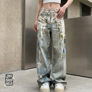 Könnte beinhalten: Hellblaue Jeans mit dekorativen Verzierungen. Die Jeans hat ein weites Bein-Design mit weißen und blauen Akzenten und Perlenketten. Die Jeans haben einen Used-Look und werden mit weißen Turnschuhen kombiniert. Das Logo "JAY DRIP HAUS" ist sichtbar.