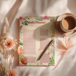Könnte beinhalten: Ein täglicher Planer mit floralem Thema mit dem Text "Daily Planner" und Abschnitten für Zeitplan, Erinnerung und To-Do-Liste. Ein Stift liegt auf dem Planer, neben einer Tasse Kaffee auf einer Untertasse. Der Planer ist mit rosa und grünen floralen Designs verziert.