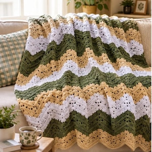 Könnte beinhalten: Eine Häkeldecke mit Zickzackmuster und abwechselnden horizontalen Streifen in Olivgrün, Creme und Weiß. Das Textil hat ein strukturiertes Lochmuster mit einem gewellten Rand und liegt über einem Sofakissen.