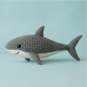 Könnte beinhalten: Ein gehäkeltes Hai-Spielzeug in Grau und Weiß, mit einem lächelnden Gesicht und schwarzen Augen. Der Hai hat eine Rückenflosse, Brustflossen und eine Schwanzflosse. Das Spielzeug ist etwa 30 cm lang und vor einem hellblauen Hintergrund.