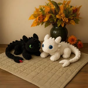 May include: Two crocheted dragon plushies, one black with green eyes and a red tongue, the other white with blue eyes. They are lying on a tan quilted surface, with a vase of fall foliage in the background.