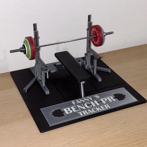 Könnte beinhalten: Ein Miniatur-Bankdrück-Set mit einer schwarzen Bank, silbernen Stützen und einer Langhantel mit bunten Gewichtsscheiben. Das Set enthält eine schwarze Basis mit dem Text "FANNY'S BENCH PR TRACKER".