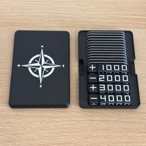 Può includere: Un componente di gioco rettangolare nero con un design a rosa dei venti bianca con le marcature N, S, E e W. Accanto, un vassoio nero contiene una pila di tessere nere con testo bianco che mostra segni più e meno seguiti dai numeri da 1000 a 4000.