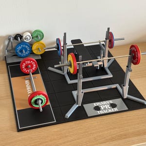 May include: A miniature weightlifting set with a bench press, squat rack, and barbell with colorful weight plates. The set includes a rack of additional weights and a platform with the text "MINIMAX" and "EINARS'S PR TRACKER".