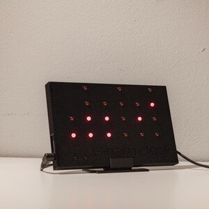 Pode incluir: Um dispositivo eletr&oacute;nico retangular preto com uma grelha de pequenas luzes circulares, algumas das quais iluminadas a vermelho. O dispositivo possui um suporte, um cabo de alimenta&ccedil;&atilde;o lateral e o texto "every clock" em relevo.