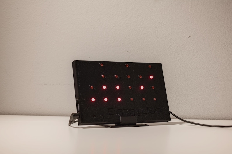 Pode incluir: Um dispositivo eletr&oacute;nico retangular preto com uma grelha de pequenas luzes circulares, algumas das quais iluminadas a vermelho. O dispositivo possui um suporte, um cabo de alimenta&ccedil;&atilde;o lateral e o texto "every clock" em relevo.