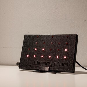 Pode incluir: Um rel&oacute;gio bin&aacute;rio retangular preto com superf&iacute;cie texturizada e suporte. O dispositivo possui v&aacute;rias pequenas luzes LED vermelhas circulares dispostas em linhas e colunas. O texto "Binary clock" est&aacute; gravado na parte inferior da caixa.