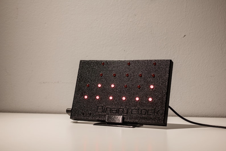 Pode incluir: Um rel&oacute;gio bin&aacute;rio retangular preto com superf&iacute;cie texturizada e suporte. O dispositivo possui v&aacute;rias pequenas luzes LED vermelhas circulares dispostas em linhas e colunas. O texto "Binary clock" est&aacute; gravado na parte inferior da caixa.