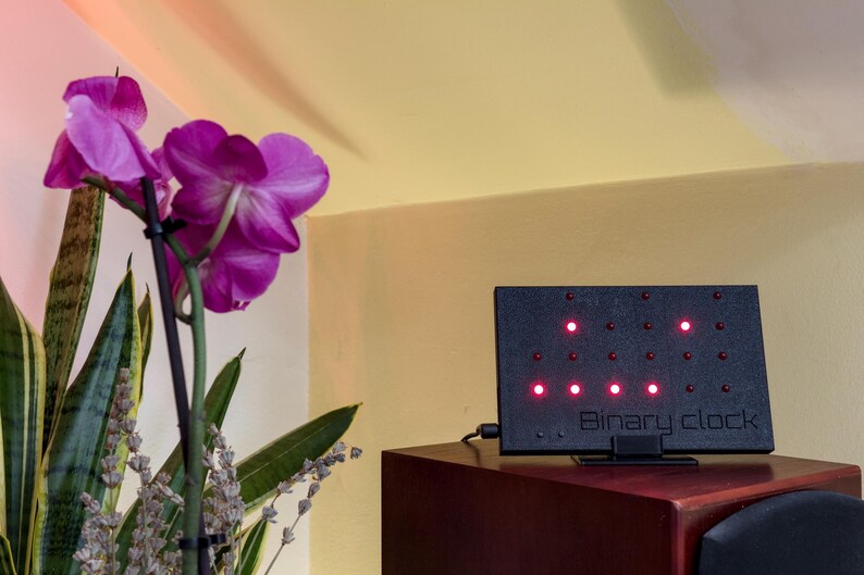 Pode incluir: Um rel&oacute;gio bin&aacute;rio eletr&oacute;nico retangular preto com uma grelha de pequenas luzes LED vermelhas e o texto "Binary clock" em relevo na parte frontal. O dispositivo est&aacute; posicionado sobre uma superf&iacute;cie de madeira escura junto a uma orqu&iacute;dea.