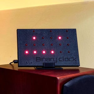 May include: A rectangular black binary clock with a textured surface and a stand. The front panel features a grid of small circular red LED lights, some of which are illuminated. The text "Binary clock" is embossed in the lower center of the device.