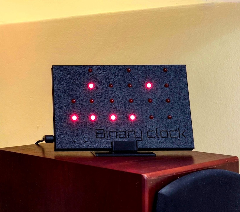 Pode incluir: Um rel&oacute;gio bin&aacute;rio retangular preto com superf&iacute;cie texturizada e suporte. O painel frontal apresenta uma grade de pequenas luzes LED vermelhas circulares, algumas das quais est&atilde;o acesas. O texto "Binary clock" est&aacute; gravado no centro inferior.