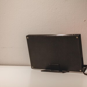 Pode incluir: Um dispositivo eletr&oacute;nico retangular preto com acabamento texturizado, fixado por quatro parafusos prateados nos cantos. O dispositivo est&aacute; montado num suporte preto e possui um cabo de alimenta&ccedil;&atilde;o preto que se estende pelo lado direito.