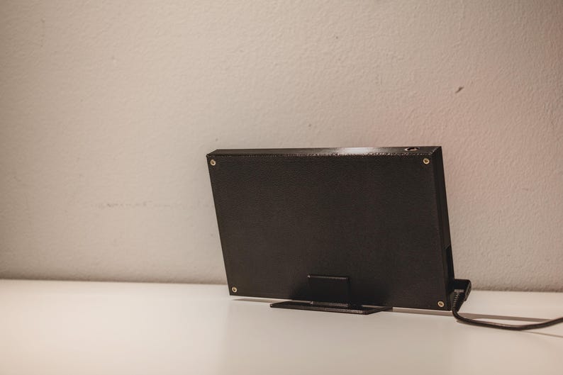 Pode incluir: Um dispositivo eletr&oacute;nico retangular preto com acabamento texturizado, fixado por quatro parafusos prateados nos cantos. O dispositivo est&aacute; montado num suporte preto e possui um cabo de alimenta&ccedil;&atilde;o preto que se estende pelo lado direito.