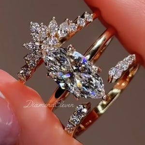 May include: A set of three gold rings with sparkling diamond accents. The central ring features a large marquise-cut diamond. The other two rings have smaller diamonds. The rings are held up by a person's fingers.