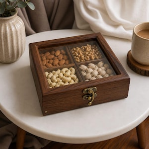 Op de afbeelding: Een houten notenserveerdoos met een glazen bovenkant, verdeeld in vier secties. Elke sectie is gevuld met een ander type noot: amandelen, walnoten, cashewnoten en pistachenoten. De doos is afgesloten met een messing sluiting.
