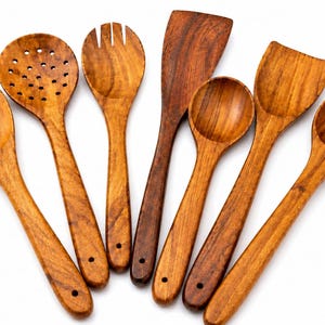 Può includere: Un set di sette utensili da cucina in legno disposti a ventaglio, tra cui un cucchiaio forato, una forchetta, una spatola piatta e quattro cucchiai rotondi di varie dimensioni, con venature del legno marrone e un foro all'estremità del manico.