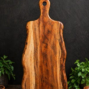 May include: A wooden cutting board with a handle and a hole for hanging. The board has a natural wood grain pattern with shades of brown and tan. The edges have a wavy, organic shape.