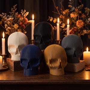 Könnte beinhalten: Eine Sammlung von fünf Totenkopfskulpturen mit vertikaler Rippenstruktur. Die Totenköpfe sind in Weiß, Schwarz, Dunkelblau, Senfgelb und Dunkelgrau gehalten. Jeder Totenkopf steht auf einem kleinen Holzblock inmitten brennender Stabkerzen.