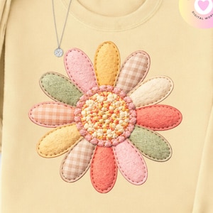 Puede incluir: Sudadera amarilla clara con un gran diseño de margarita colorida. La margarita tiene pétalos en varios colores pastel y un centro texturizado. Se ve un collar plateado con un pequeño colgante cerca del cuello. El diseño recuerda a un patchwork.