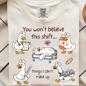 Op de afbeelding: Een crèmekleurig t-shirt met een afbeelding van cartoon-eenden verkleed als medisch personeel. De tekst luidt: "You won't believe this shift... things I can't make up." De eenden zijn afgebeeld met een stethoscoop, klembord, medicijnfles, thermometer en een dienblad met benodigdheden, waarbij één eend op een ziekenhuisbed ligt.