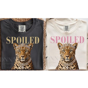 Op de afbeelding: Twee t-shirts, één antracietgrijs en één crème, elk met de tekst SPOILED in dikke hoofdletters boven een afbeelding van een luipaard met een met juwelen bezette kroon en een dubbele parelketting met een centrale druppelvormige hanger.