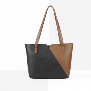 Könnte beinhalten: Eine Tragetasche mit einem farblich abgesetzten Design, bestehend aus einer schwarzen linken Seite und einer braunen rechten Seite, die durch eine diagonale Naht getrennt sind. Die Tasche hat zwei lange, schmale braune Schulterriemen.