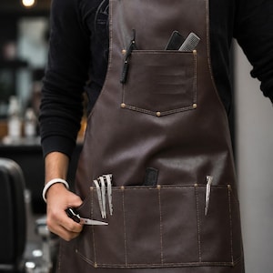 May include: A dark brown apron featuring a chest pocket containing two black combs and a straight razor, plus a lower section with multiple pockets holding silver hair clips and a black comb. The apron has metal hardware and visible stitching.