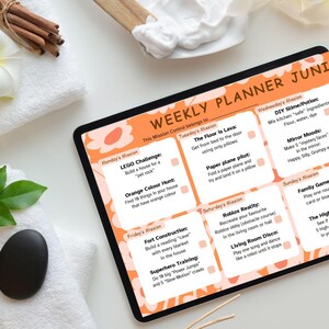 May include: A black tablet with an orange border shows a weekly planner for children. The planner features daily activities such as building a fort, playing Roblox, and making slime. The background includes white towels, a black stone, and wooden sticks.