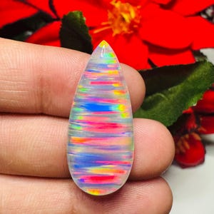 May include: A teardrop-shaped, iridescent gemstone with a rainbow of colors, including red, blue, yellow, and green. The stone is held in a hand, with a red flower in the background. The gemstone is likely an opal.