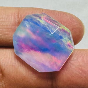 May include: A faceted hexagonal gemstone displaying an iridescent play of color with translucent blue, violet, and pink hues. The stone features a smooth, polished surface with internal light reflections and a distinct geometric shape.
