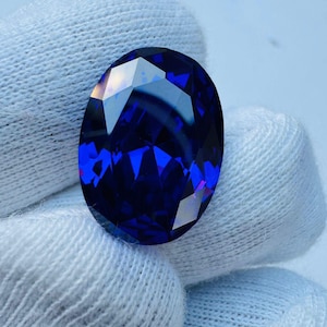 May include: A faceted, oval-shaped, deep blue gemstone. The gem is held by a white fabric, and the facets reflect light, creating a brilliant display of color and sparkle. The gemstone's rich color is a striking feature.